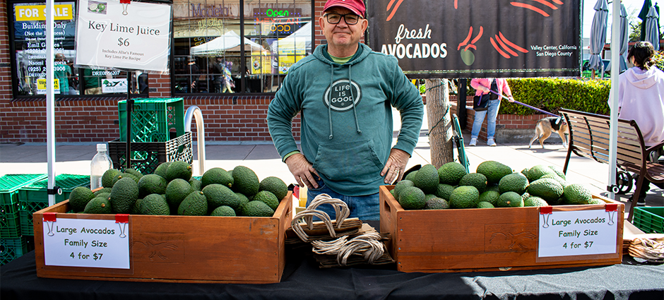 Photo of producer standing by avocados in booth at market